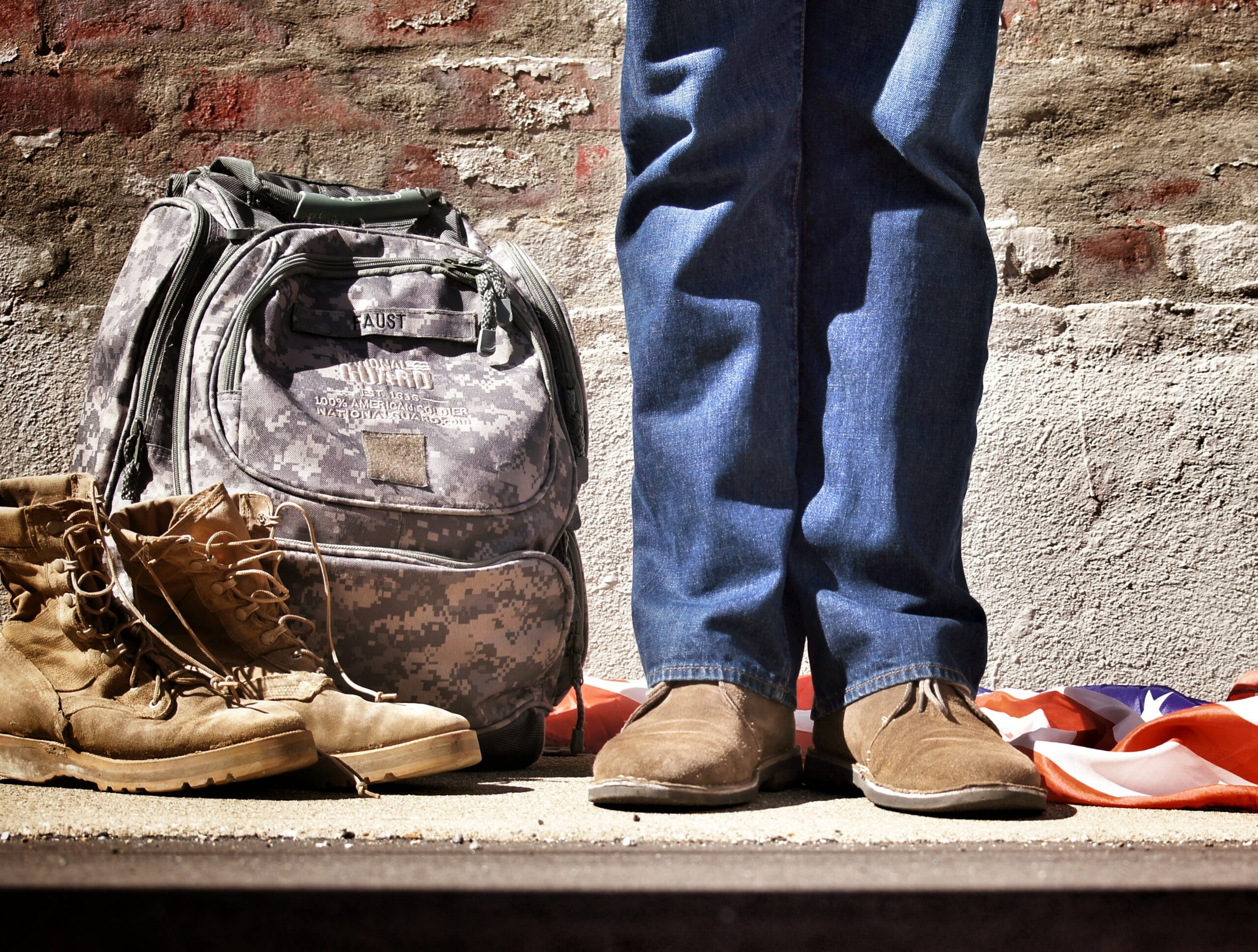 The legs of a man in jeans next to a camouflage rucksack and military boots.